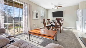 A spacious living room with a sliding glass door to a patio, a dining area with wooden chairs, a flat-screen TV mounted on the wall, and a refrigerator in the kitchen area. - Windsor Cottage Fall Creek Beauty by Table Rock! (Branson)