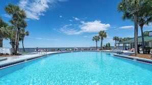 A clear blue pool with palm trees, a covered seating area, and a view of the ocean. - Seashell Shuffle at Pilot House (Miramar Beach)