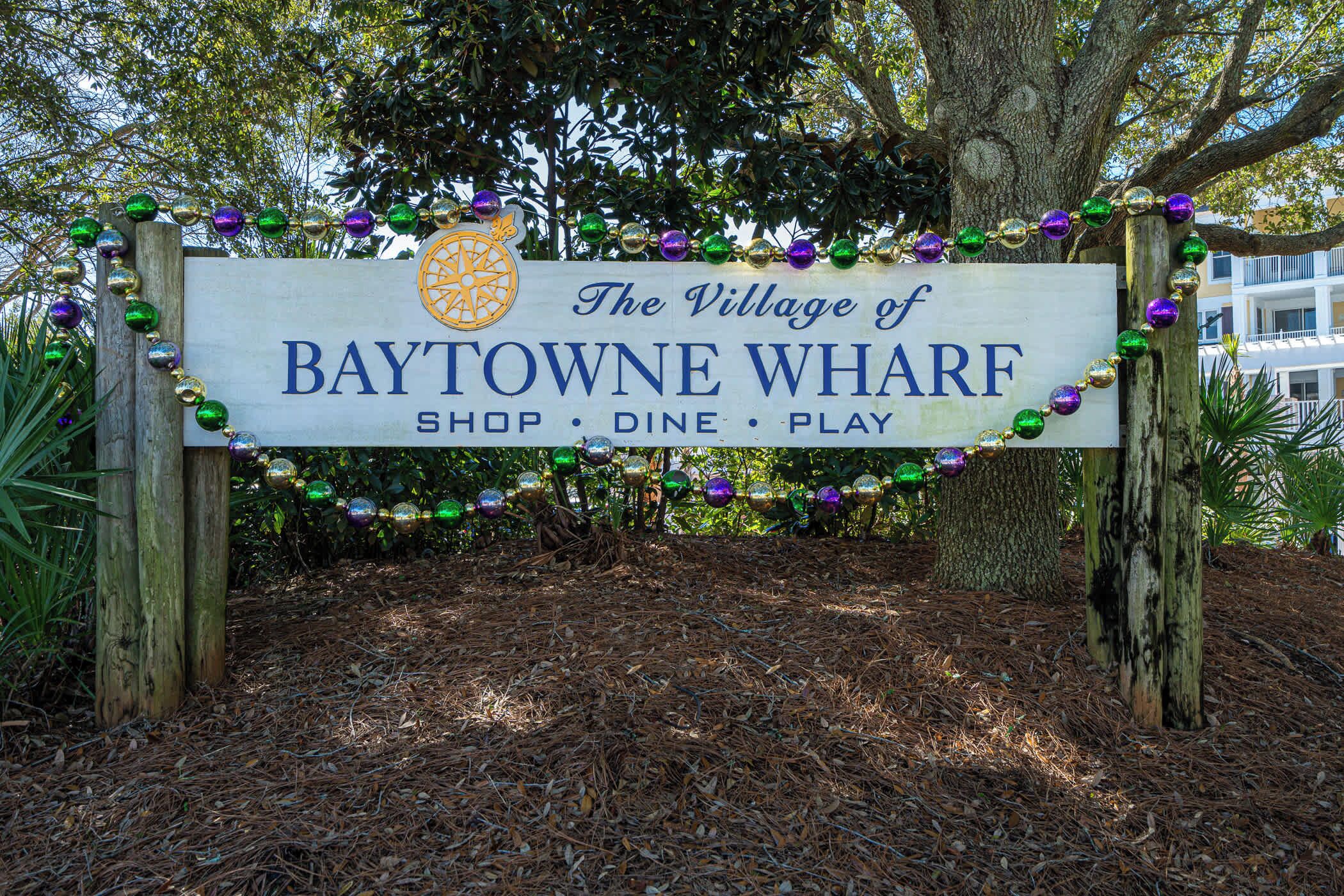 A wooden sign with the text "The Village of BAYTOWNE WHARF SHOP • DINE • PLAY" surrounded by decorative beads.