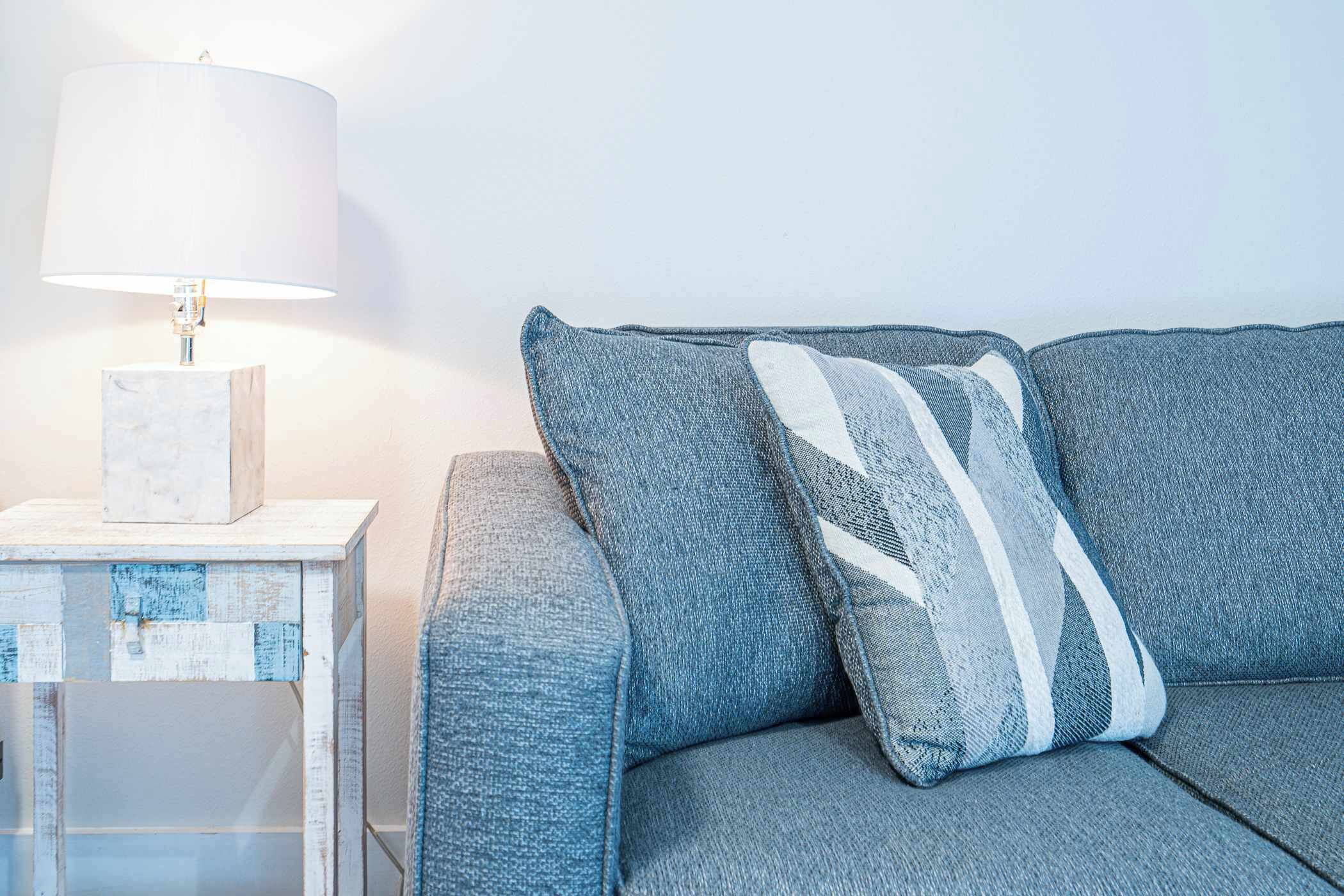 A sofa with a grey fabric and white stripes pillow, a white side table with a lamp, and a white block on the table.