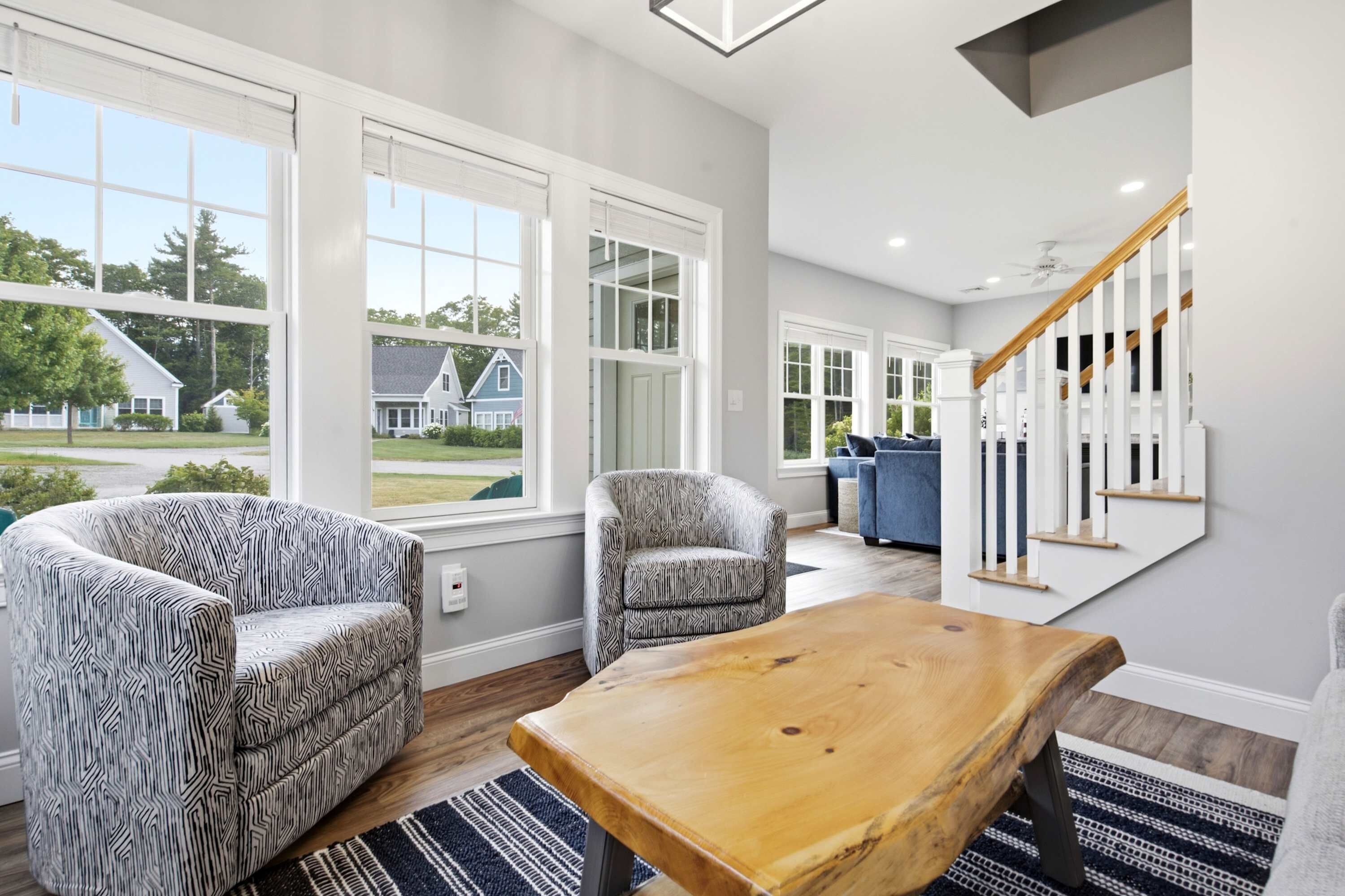 A spacious living room with large windows, a wooden coffee table, two patterned armchairs, and a staircase with white railings.