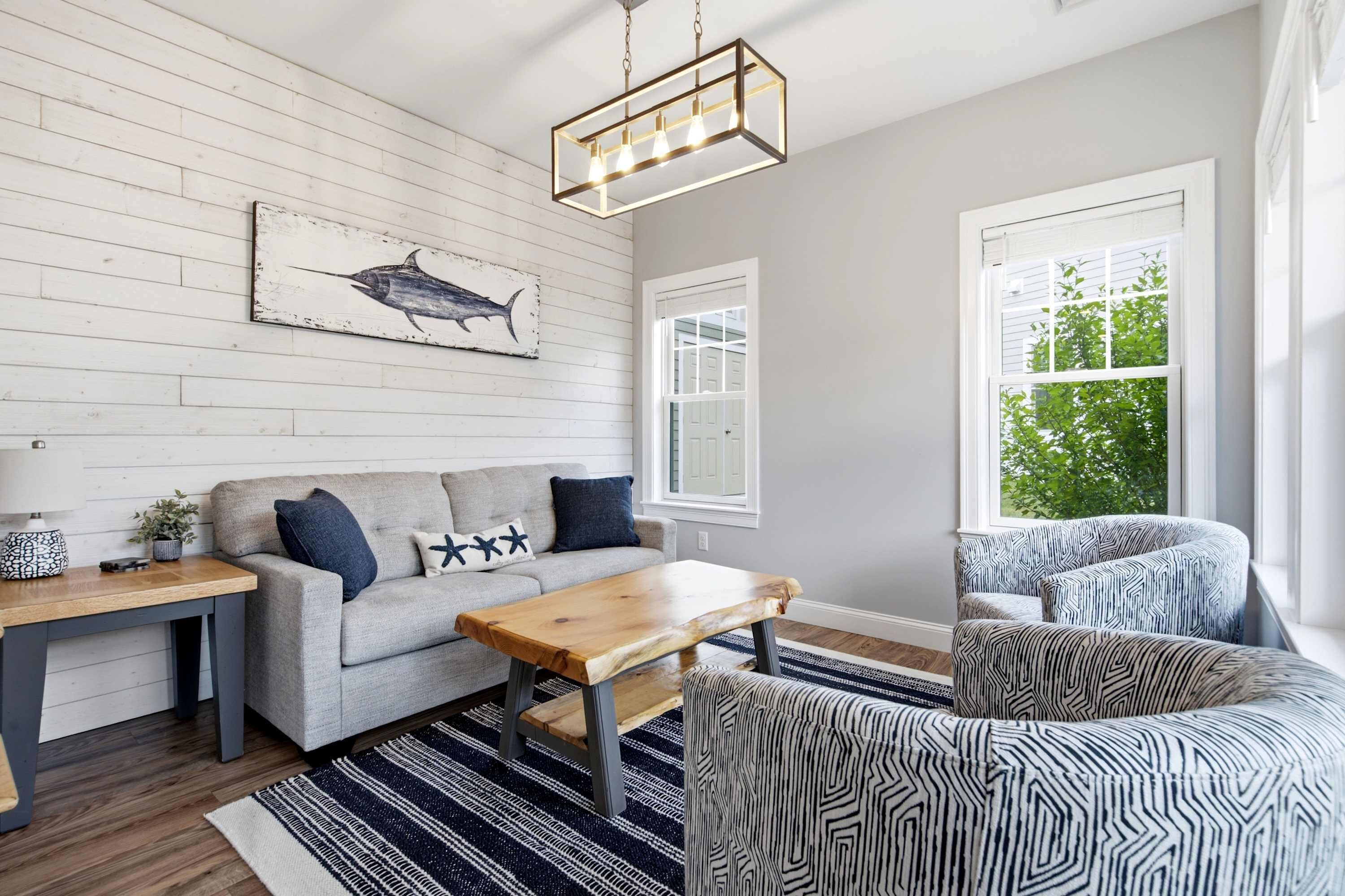 A living room with a grey sofa, a wooden coffee table, a patterned rug, a wall-mounted light fixture, and a window with a view of greenery.