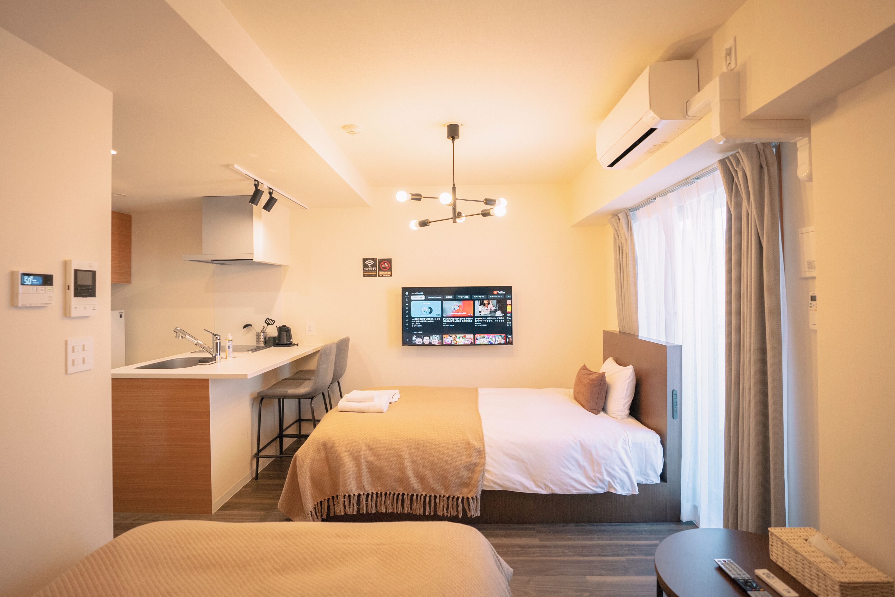 A hotel room with a kitchenette, a bed with a headboard, a desk with a chair, a TV mounted on the wall, and a window with curtains.