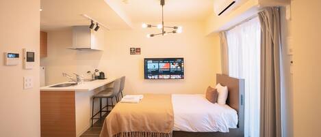 A hotel room with a kitchenette, a bed with a headboard, a desk with a chair, a TV mounted on the wall, and a window with curtains.