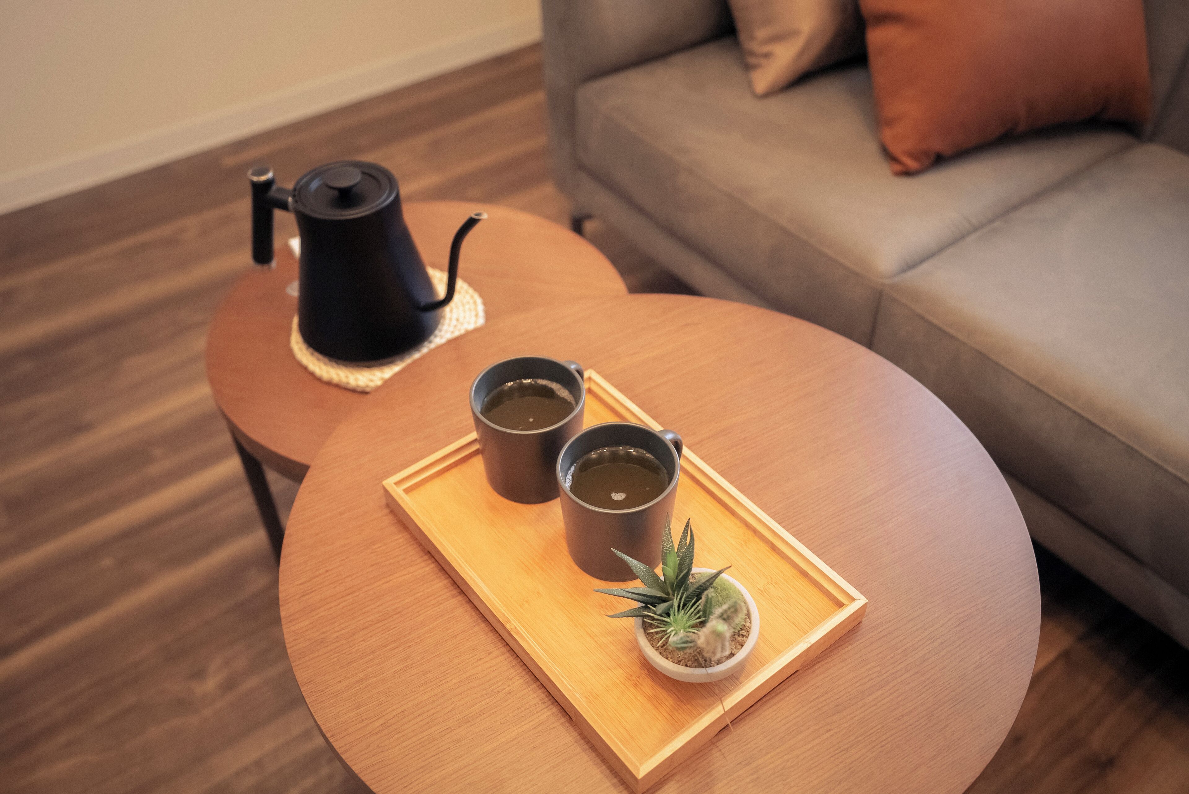 A round wooden table with a tray holding two cups of tea, a small potted plant, and a black teapot.