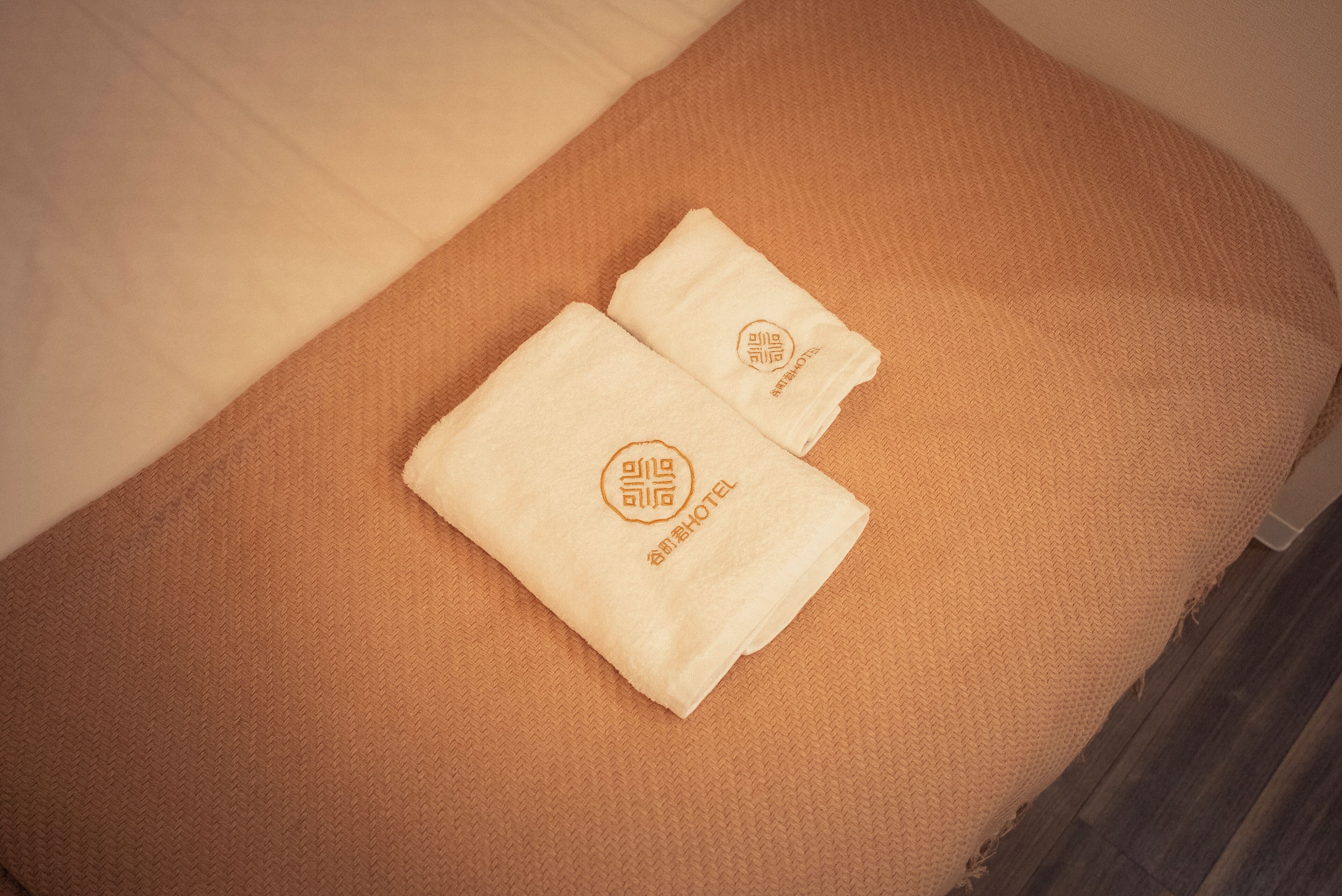 A neatly made bed with a beige blanket and two folded towels with a logo and text on them.