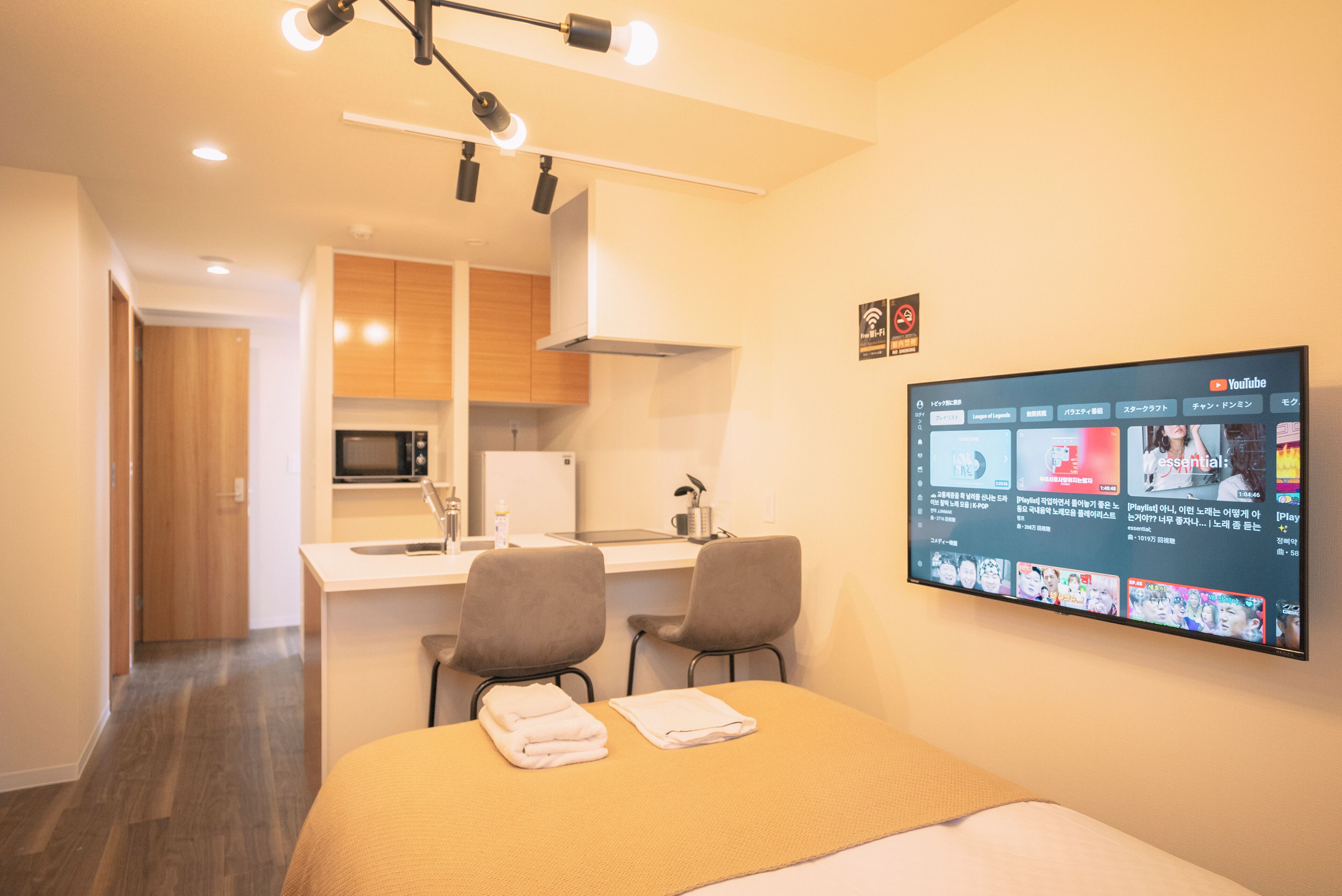 A modern hotel room with a bed, a desk with two chairs, a flat-screen TV, and a kitchenette.