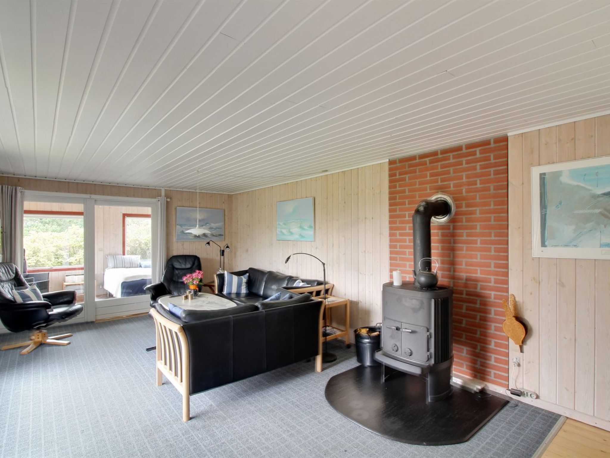 Une pièce chaleureuse avec un canapé en cuir noir, une table basse en bois, une chaise noire, une petite table avec une lampe et un mur en briques avec un poêle à bois.