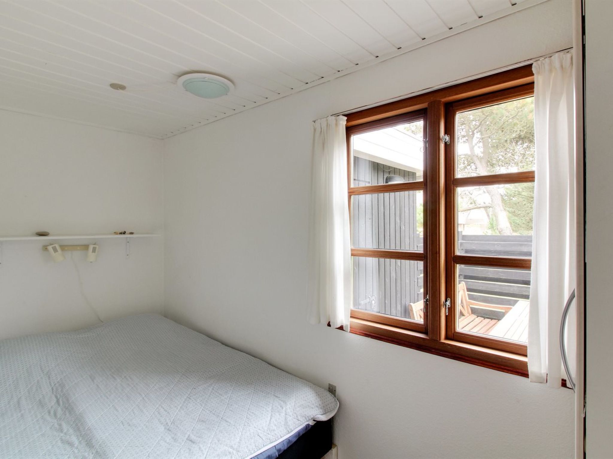 Una habitación pequeña con cama individual, ventana de madera, paredes blancas y una lámpara en el techo.