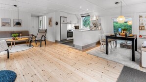 Amplia sala de estar con suelo de madera, un sofá, una mesa de comedor y una cocina con armarios blancos y un ventanal grande.