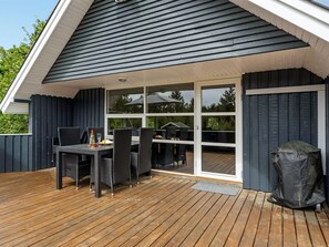 Eine überdachte Terrasse im Freien mit Tisch und Stühlen, einem Grill und einer Schiebetür zu einer Holzterrasse.