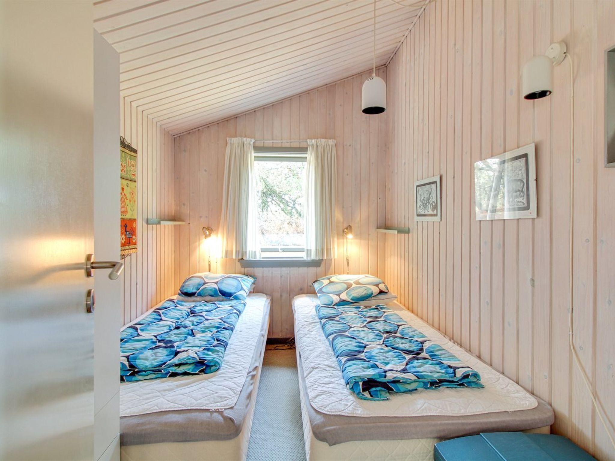 A cozy room with two beds, wooden walls, a window with curtains, and wall-mounted lights.