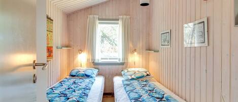 A cozy room with two beds, wooden walls, a window with curtains, and wall-mounted lights.