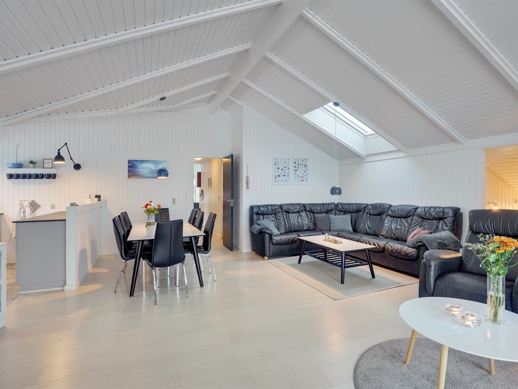 A spacious loft with a high ceiling, a dining area with a table and chairs, a black leather sofa, and a small round table with a vase of flowers.