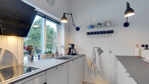 Une cuisine moderne avec des meubles blancs, un four encastré, un évier et une fenêtre qui laisse entrer la lumière naturelle. On y trouve deux lampes suspendues, une étagère avec des tasses bleues et une horloge au mur.