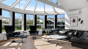 Ein geräumiger Raum mit hoher Decke, großen Fenstern, einem schwarzen Ledersofa, einem runden Couchtisch und einem schwarzen Sessel.