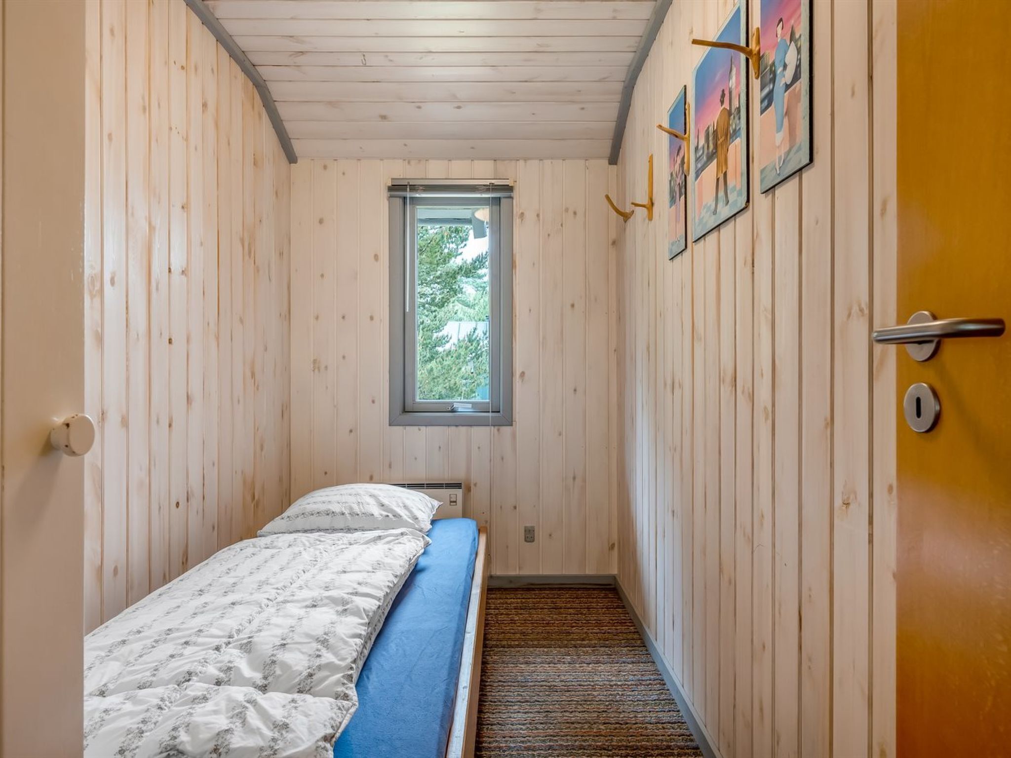 A narrow room with wooden walls, a single bed with a blue mattress, a window with a view of trees, and a door with a handle.