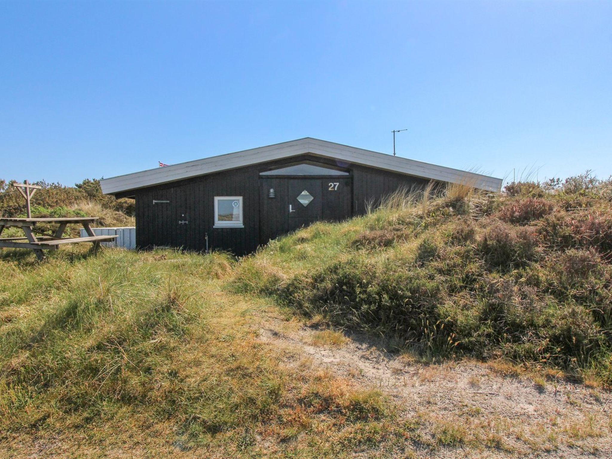 Un petit bâtiment en bois sombre, portant le numéro « 27 » sur le côté, situé sur une colline herbeuse, avec une table de pique-nique à proximité.