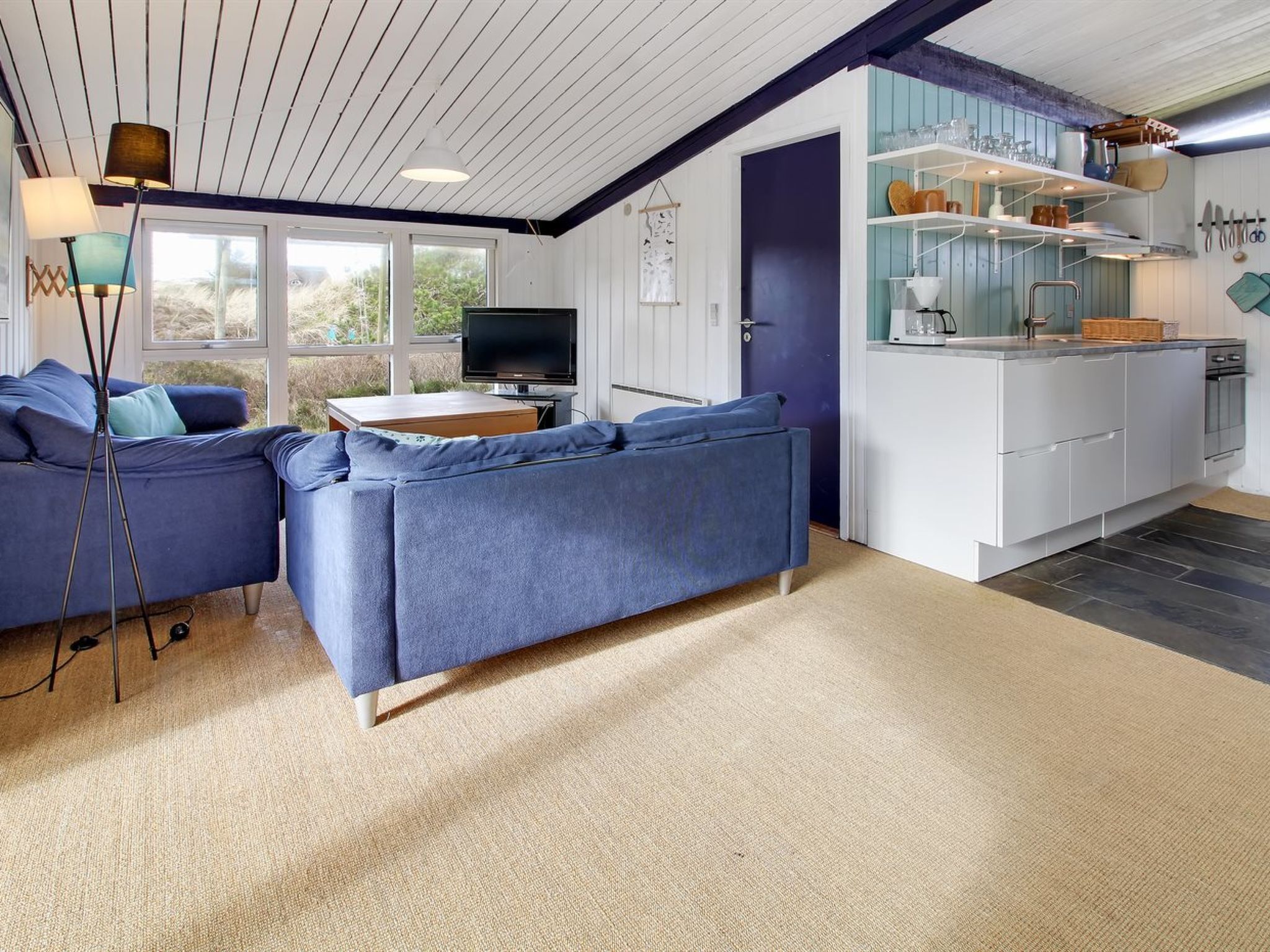 Un salon moderne avec un canapé bleu, un téléviseur et un coin cuisine doté de meubles blancs et de touches de couleur bleue.