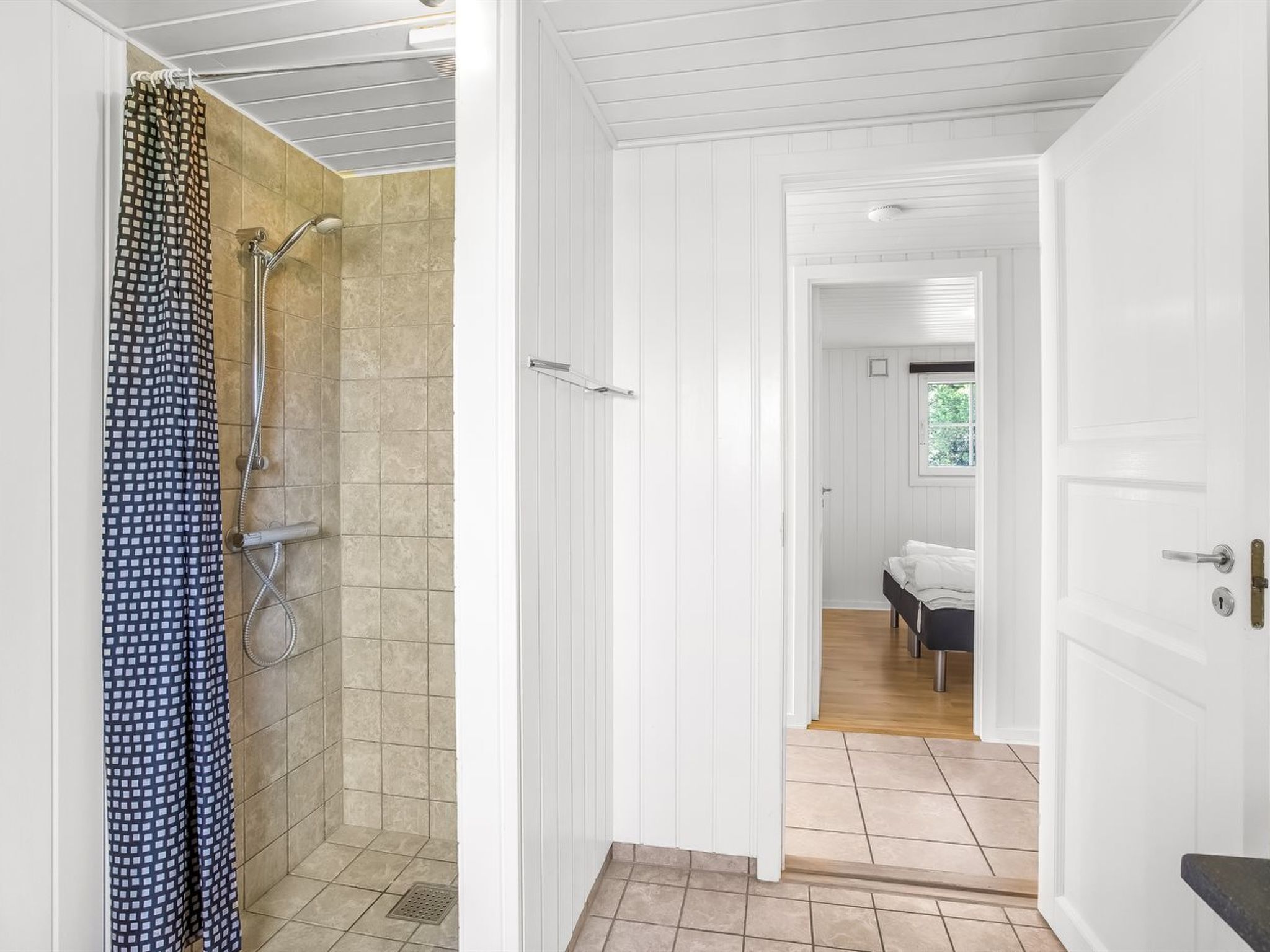 A bathroom with a shower, a toilet, and a sink. There is a door leading to a bedroom with a bed and a window.