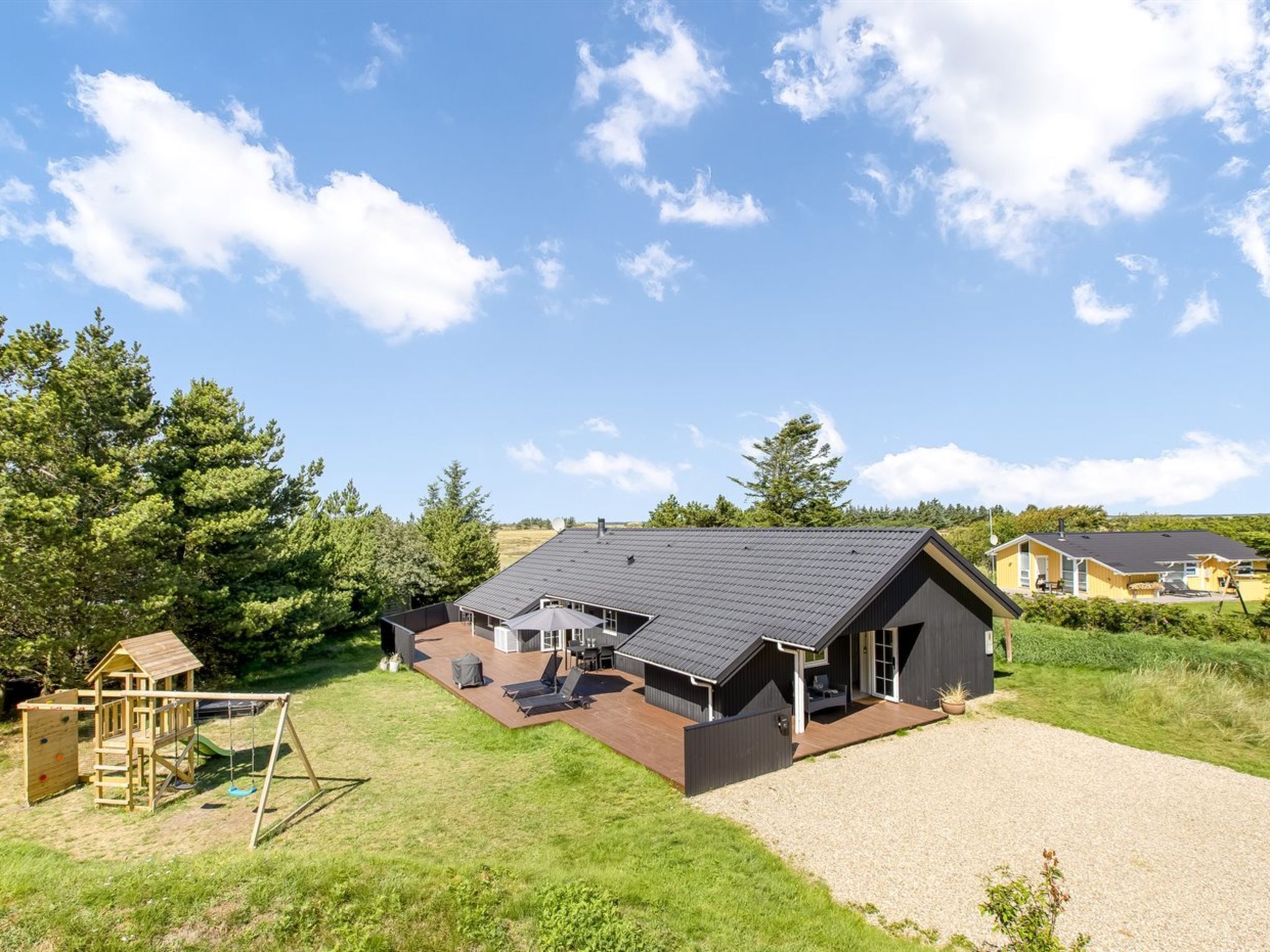 A modern house with a large deck, a play area, and a gravel driveway.