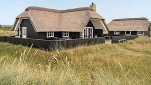 Dos casas rurales con techos de paja, paredes negras y ventanas blancas, rodeadas de hierba alta.