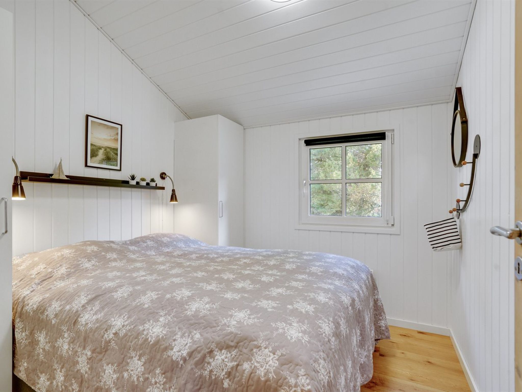 A bedroom with a bed, a window, a wall-mounted coat rack, and a shelf with plants.