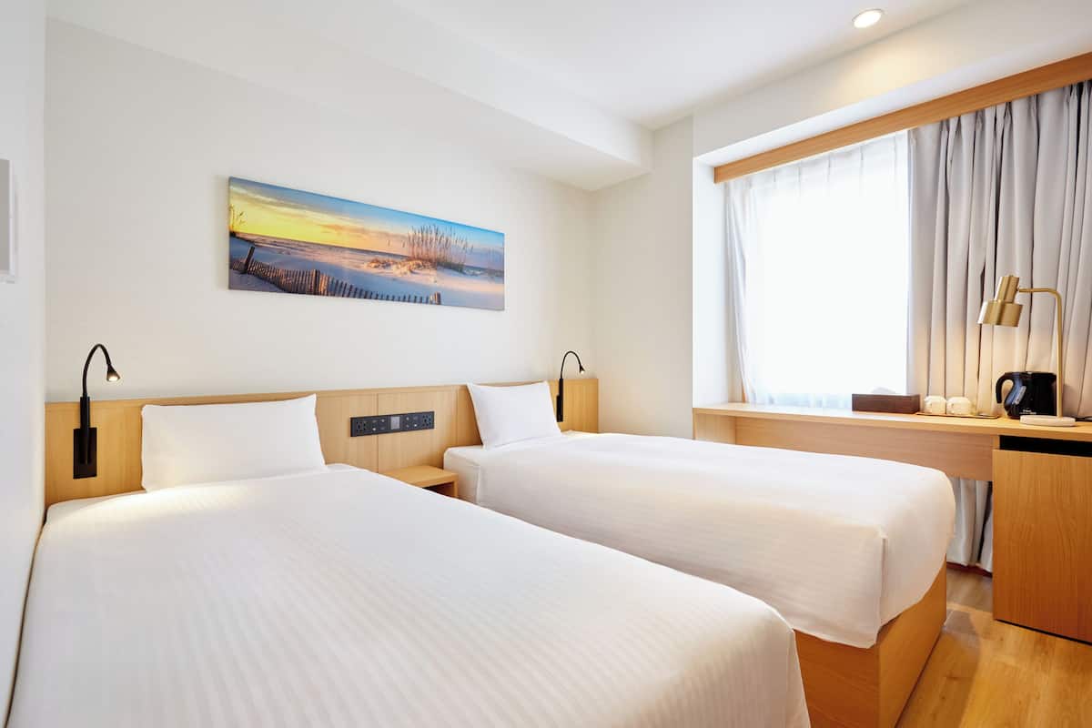 Habitación de hotel con dos camas, un cuadro de una puesta de sol, un escritorio con lámpara y una ventana con cortinas.
