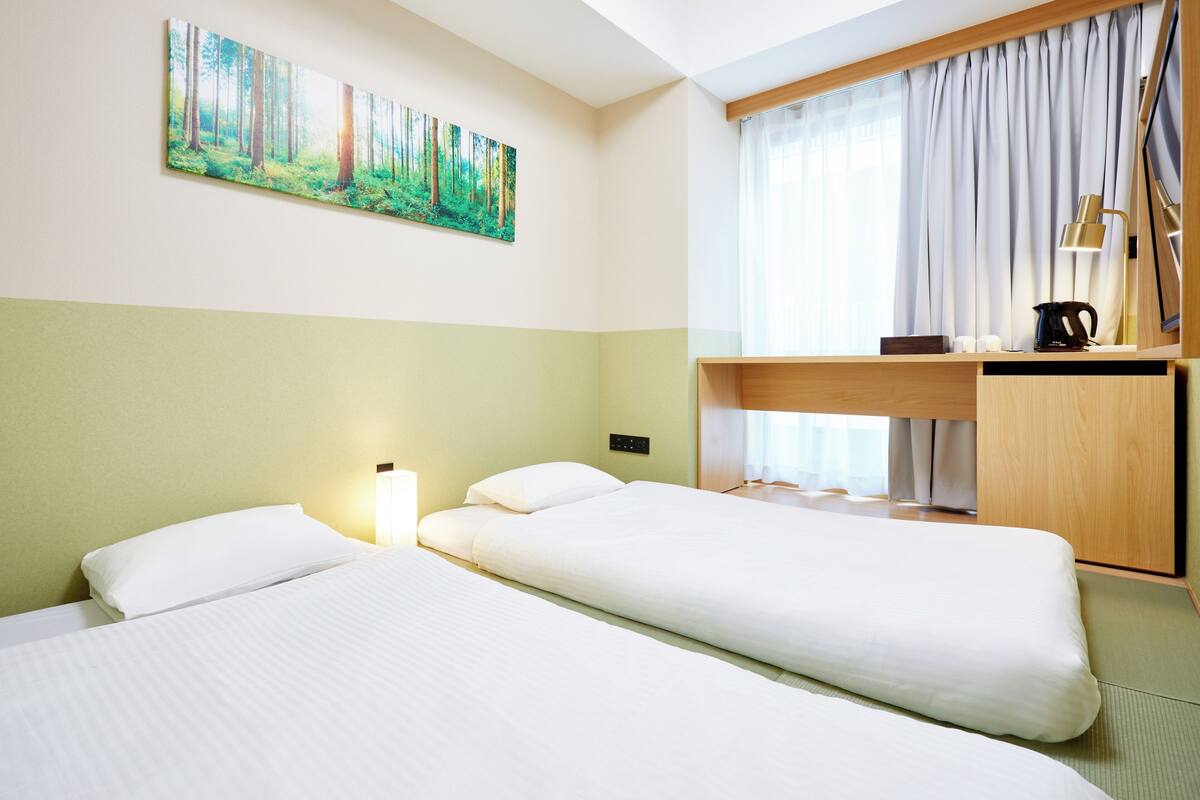 Habitación de hotel con dos camas, un cuadro de un bosque, un escritorio con una lámpara y una ventana con cortinas.