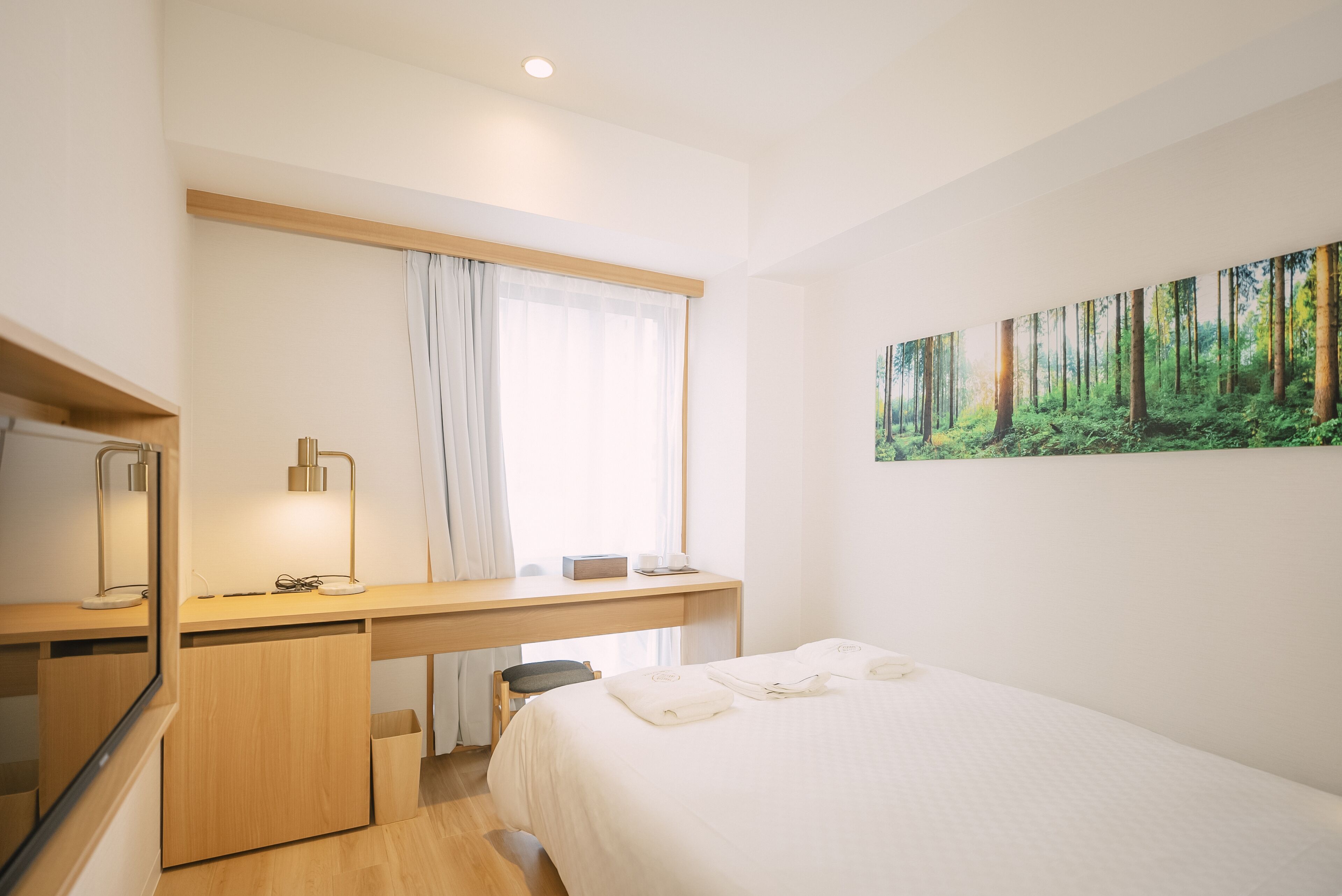 A modern hotel room with a wooden desk, a bed with neatly folded towels, a large forest-themed painting on the wall, and a window with sheer curtains.