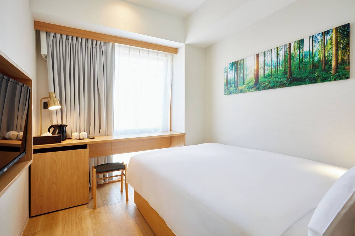 Habitación de hotel con cama, escritorio y silla, ventana con cortinas, un cuadro de un bosque y una lámpara.