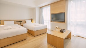 Une chambre d’hôtel avec deux lits, un bureau en bois, une télévision et de grandes fenêtres avec des rideaux légers.