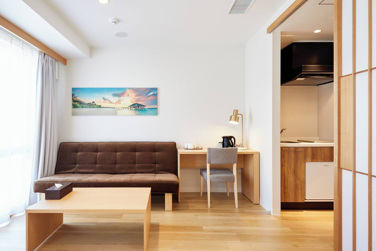 Una sala de estar moderna con un sofá, un escritorio y una silla. Hay un cuadro en la pared y se ve un área de cocina a través de una puerta abierta.