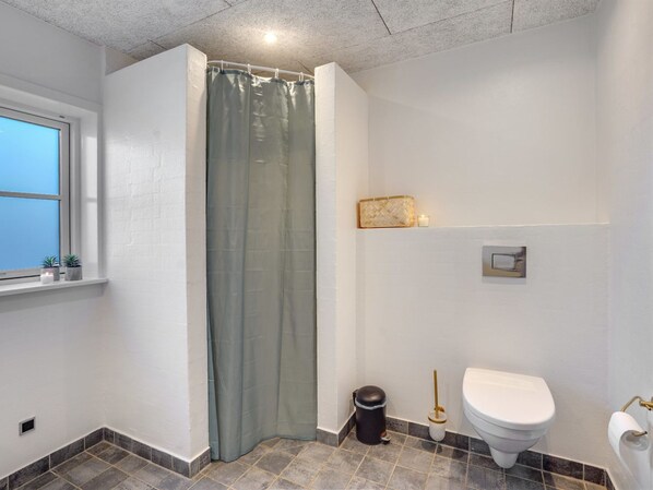 A bathroom with a shower curtain, a toilet, a trash bin, a small shelf with a candle, and a window with a view.