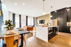 The kitchen island, dining table, and living room form one open space so nobody cooking misses the group. Neon signs glow overhead and the whole house connects in sight lines