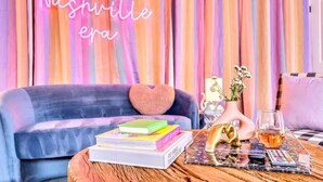 Een gezellige woonkamer met een neonbord waarop staat 'In my Nashville era', een blauwe bank met een hartvormig kussen, een bijzettafeltje met boeken, een vaas met bloemen en een glas whisky.