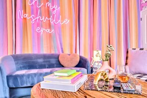 A cozy living room with a neon sign reading "In my Nashville era," a blue sofa with a heart-shaped pillow, a coffee table with books, a vase with flowers, and a glass of whiskey.