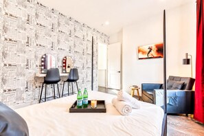 Una habitación de hotel con una cama, una zona de estar con sillas, una mesita con botellas y vasos, un cuadro en la pared y una cortina roja.