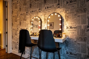 Ein Waschtisch mit Spiegeln und Beleuchtung, ein Stuhl und eine Wand mit Zeitungsmustern.
