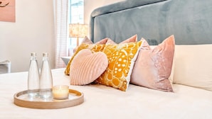 Een netjes ingerichte hotelkamer met een bed, kussens, een dienblad met flessen en een kaars, en een lamp op de achtergrond.