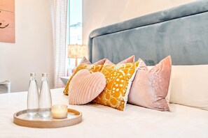 A neatly arranged hotel room with a bed, pillows, a tray with bottles and a candle, and a lamp in the background.
