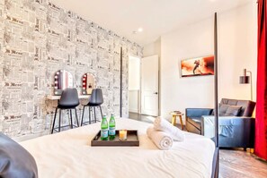 A hotel room with a bed, a desk with two chairs, a sofa, and a wall with newspaper wallpaper.