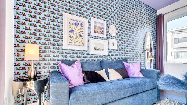 Een woonkamer met behang met een patroon, een blauwe bank met kussens, een lamp en ingelijste foto's aan de muur.