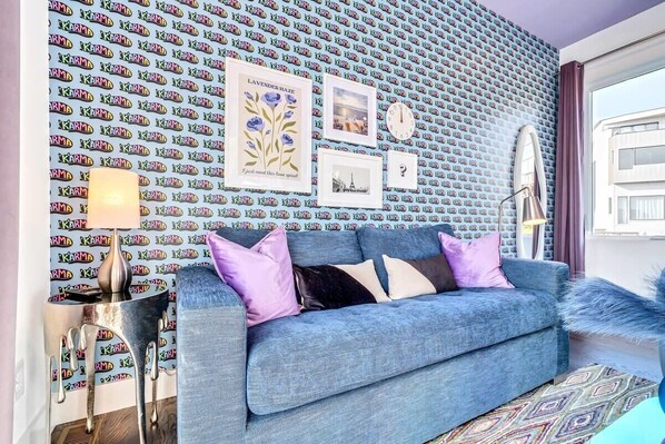 A living room with a patterned wallpaper, a blue sofa with cushions, a lamp, and framed pictures on the wall.