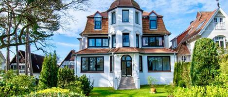 A large, multi-story house with a prominent corner turret, surrounded by well-maintained gardens and trees.