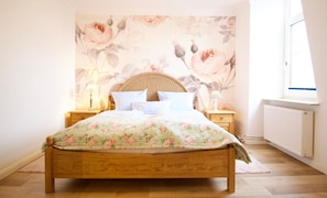 A bedroom with a wooden bed, floral wallpaper, two bedside tables with lamps, and a window with a view of the sky.