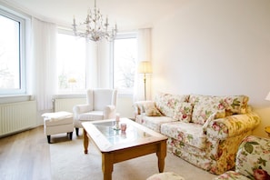 Un salón con un sofá floral, un sillón blanco, una mesa de centro de cristal y una araña.