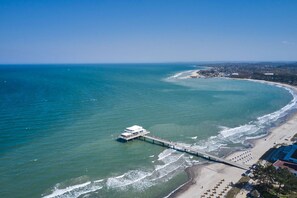 Vista aérea de uma área costeira com um cais que se estende para o mar, uma praia com espreguiçadeiras e uma linha costeira distante com edifícios.