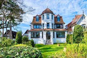 A large, multi-story house with a prominent corner turret, surrounded by well-maintained gardens and trees.