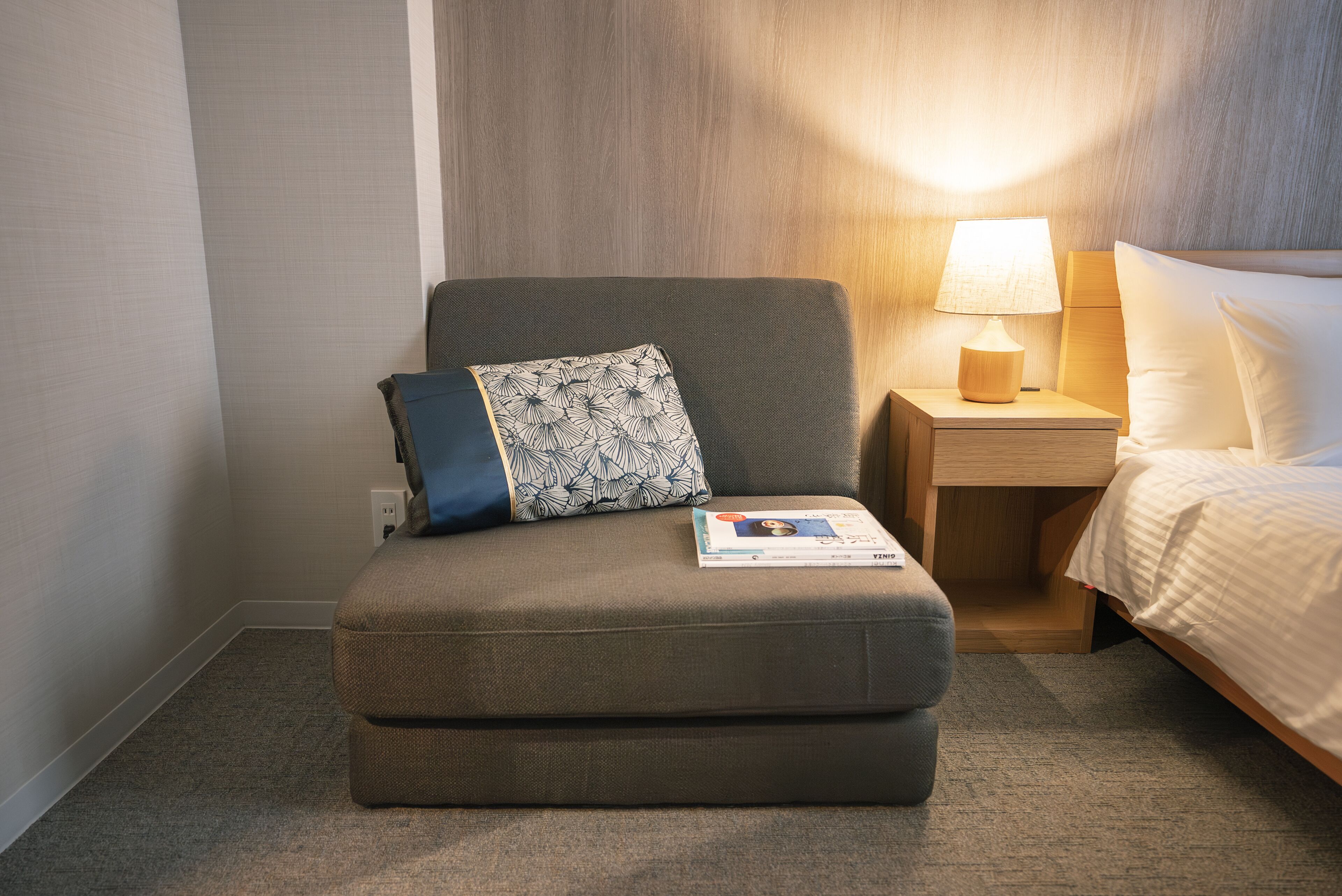 A small, dark-colored sofa with a patterned pillow and a magazine, positioned next to a wooden nightstand with a lamp.